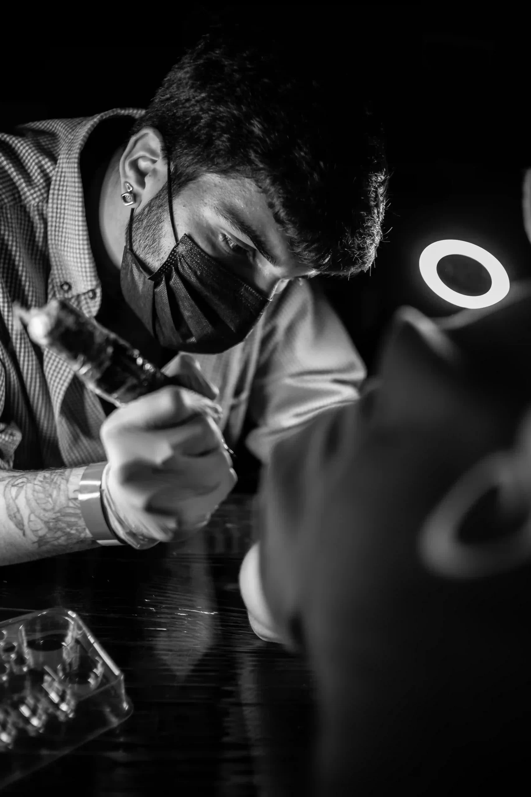 Close-up photo in black and white displaying a man wearing mask doing tattoo in another person holding a wireless tattoo machine in one hand, the background is mainly dark, but there is a ring light on the right side.